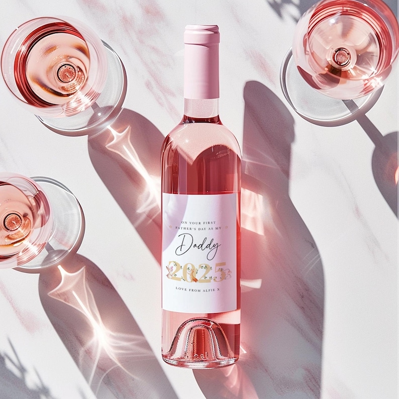 May include: A pink wine bottle with a custom label that reads "Daddy 2023" and the text "Love from Alfie S." The bottle is surrounded by three wine glasses filled with pink wine. The image is shot from above, with shadows.