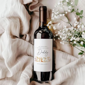 May include: A wine bottle with a custom label that reads "ON YOUR FIRST FATHER'S DAY AS MY Daddy 2025 Love From Alfie X". The bottle is filled with red wine and is set against a neutral fabric background with white flowers.