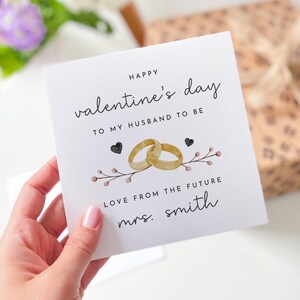 K&ouml;nnte beinhalten: Eine wei&szlig;e Karte mit schwarzem Text, der "Happy valentine's day to my husband to be Love from the future mrs. smith" lautet. Die Karte hat eine Aquarell-Illustration von zwei goldenen Ringen und einem Zweig mit rosa Beeren.