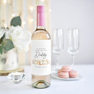 May include: A clear wine bottle with a pink cap and a white label. The label reads "ON YOUR FIRST FATHER'S DAY AS MY Daddy 2025 LOVE FROM ALFIE X". Two champagne flutes and pink macarons are in the background.