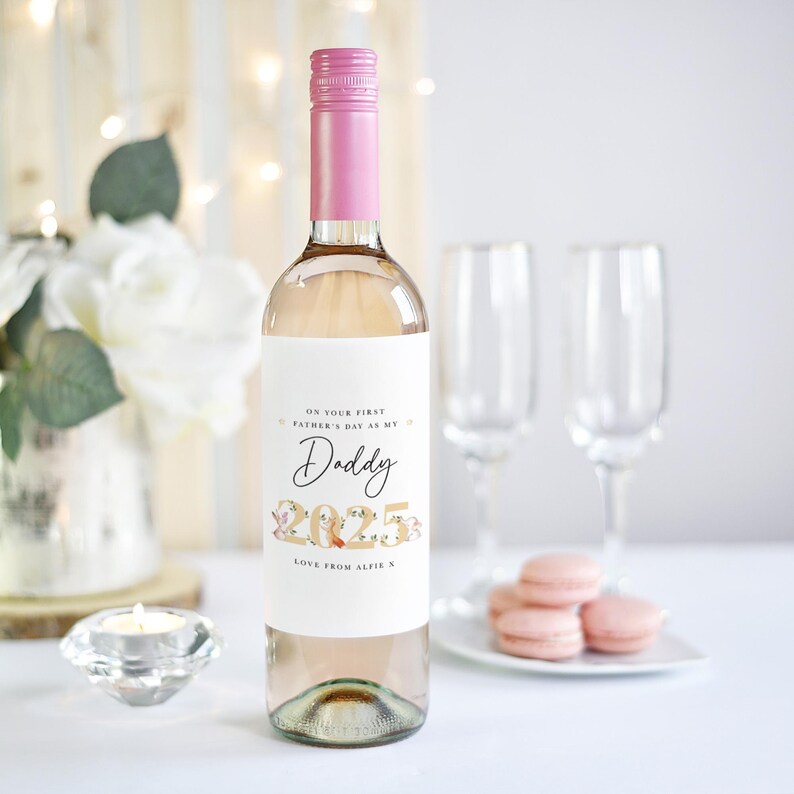 May include: A clear wine bottle with a pink cap and a white label. The label reads "ON YOUR FIRST FATHER'S DAY AS MY Daddy 2025 LOVE FROM ALFIE X". Two champagne flutes and pink macarons are in the background.