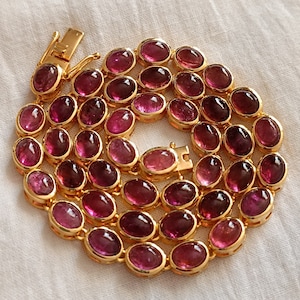 May include: A gold necklace featuring a chain of oval-shaped pink gemstones. The necklace has a clasp fastening.