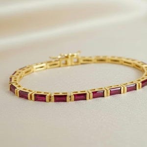 May include: A gold bracelet with rectangular, deep red gemstones. The bracelet is set in a gold frame and has a clasp closure. The jewelry is displayed on a cream-colored surface.