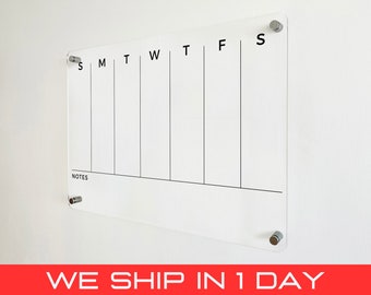 May include: A clear acrylic weekly planner with black text and lines. The days of the week are labeled across the top, with a section for notes at the bottom. The planner is mounted on a white wall with silver hardware.