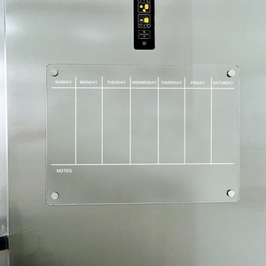 Puede incluir: Un planificador semanal de borrado en seco blanco con una secci&oacute;n de notas. El planificador est&aacute; sujeto a un refrigerador de acero inoxidable con botones magn&eacute;ticos plateados.