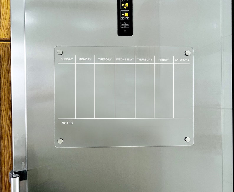Puede incluir: Un planificador semanal de borrado en seco blanco con una secci&oacute;n de notas. El planificador est&aacute; sujeto a un refrigerador de acero inoxidable con botones magn&eacute;ticos plateados.