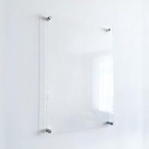 May include: A rectangular clear acrylic sign panel mounted to a wall with four silver-toned metal standoffs at each corner. The transparent board features a smooth, glossy surface and is positioned vertically, providing a clean and neutral display.