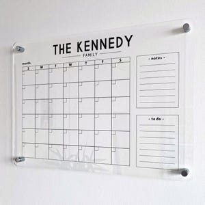 May include: A clear acrylic wall calendar featuring black printed text and grid lines. The header reads THE KENNEDY FAMILY, followed by a monthly calendar grid, a notes section, and a to-do list. It is mounted with four silver metal standoffs.