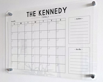 May include: A clear acrylic wall calendar featuring black printed text and grid lines. The header reads THE KENNEDY FAMILY, followed by a monthly calendar grid, a notes section, and a to-do list. It is mounted with four silver metal standoffs.