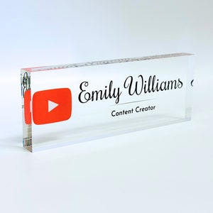 May include: A rectangular clear acrylic desk name plate featuring a red YouTube play button icon on the left side. The text Emily Williams is printed in a black script font above the words Content Creator in a simple black sans-serif font.