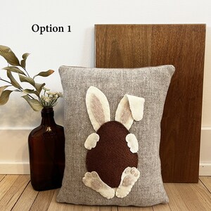 May include: A gray pillow with a brown felt bunny rabbit appliqu&eacute;. The bunny is sitting on its hind legs with its tail in the air. The bunny has pink ears and white feet.