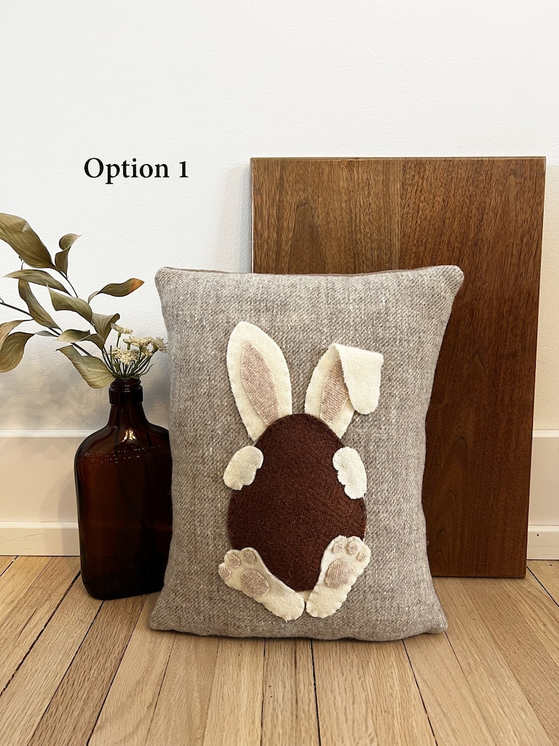 May include: A gray pillow with a brown felt bunny rabbit appliqu&eacute;. The bunny is sitting on its hind legs with its tail in the air. The bunny has pink ears and white feet.