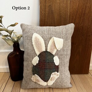 May include: A gray wool pillow with a felt bunny rabbit appliqu&eacute;. The bunny is white with pink ears and a plaid tail. The pillow is a decorative accent for a home.