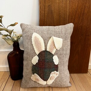 May include: A gray pillow with a felt bunny rabbit appliqu&eacute;. The bunny is sitting on its hind legs with its tail in the air. The bunny's body is a plaid fabric with a green, red, and black pattern. The bunny's ears and feet are white felt.