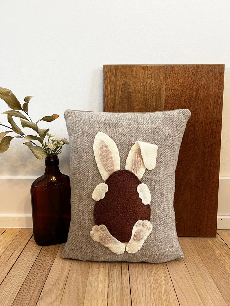 May include: A gray throw pillow with a brown felt bunny rabbit appliqu&eacute;. The bunny is sitting on its hind legs with its tail in the air. The bunny's ears are pink and its feet are white.