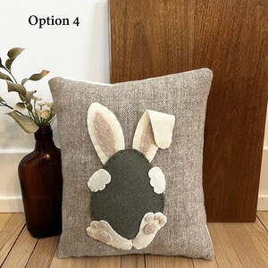 May include: A brown and beige throw pillow with a felt bunny rabbit appliqu&eacute;. The bunny is sitting on its hind legs with its tail in the air. The bunny is green and has pink ears and feet.