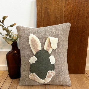 May include: A brown and beige throw pillow with a felt bunny rabbit design. The bunny is sitting on its hind legs with its tail in the air. The bunny is green and has white feet and ears.