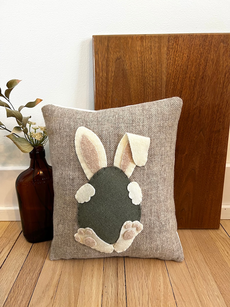 May include: A brown and beige throw pillow with a felt bunny rabbit design. The bunny is sitting on its hind legs with its tail in the air. The bunny is green and has white feet and ears.
