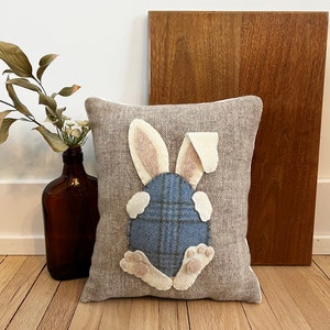 May include: A gray pillow with a white bunny rabbit appliqu&eacute;. The bunny is sitting on its hind legs with its tail tucked behind it. The bunny is wearing a blue and white plaid egg.