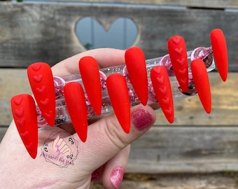 May include: Set of ten red press-on nails with a heart design. The nails are long and have a stiletto shape.