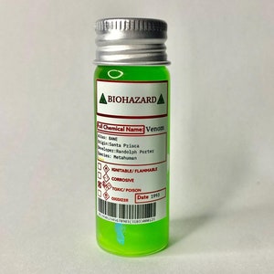 May include: A small green glass bottle with a silver lid. The bottle has a white label with the words "BIOHAZARD", "Full Chemical Name: Venom", "Alias: BANE", "Origin: Santa Prisca", "Developer: Randolph Porter", "Species: Metahuman", "Ignitable/Flammable", "Corrosive", "Toxic/Poison", and "Date 1993".