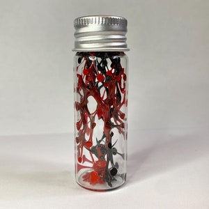 May include: A small glass bottle with a silver lid filled with red and black paint splatter.