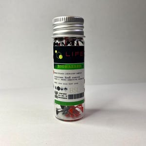 K&ouml;nnte beinhalten: Ein kleines Glasfl&auml;schchen mit einem silbernen Deckel, das rote und schwarze Plastikteile enth&auml;lt. Das Fl&auml;schchen hat ein gr&uuml;n-schwarzes Etikett mit der Aufschrift "LIFE BIOHAZARD" und dem Text "VRASITOLOGY LABORATORY SAMPLE", "VOSPECIMEN: Knull SYMBIOTE", "CASE 1: NEURO-SENSITIVE ORGANISM", "税込: 2000 C210 4197 4998", "BSL-8", "4794-012", "24-05-12", "09729826".