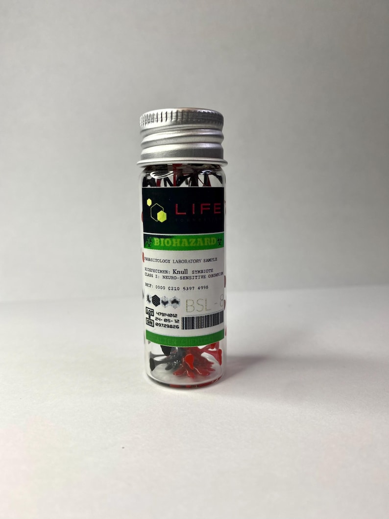 K&ouml;nnte beinhalten: Ein kleines Glasfl&auml;schchen mit einem silbernen Deckel, das rote und schwarze Plastikteile enth&auml;lt. Das Fl&auml;schchen hat ein gr&uuml;n-schwarzes Etikett mit der Aufschrift "LIFE BIOHAZARD" und dem Text "VRASITOLOGY LABORATORY SAMPLE", "VOSPECIMEN: Knull SYMBIOTE", "CASE 1: NEURO-SENSITIVE ORGANISM", "税込: 2000 C210 4197 4998", "BSL-8", "4794-012", "24-05-12", "09729826".