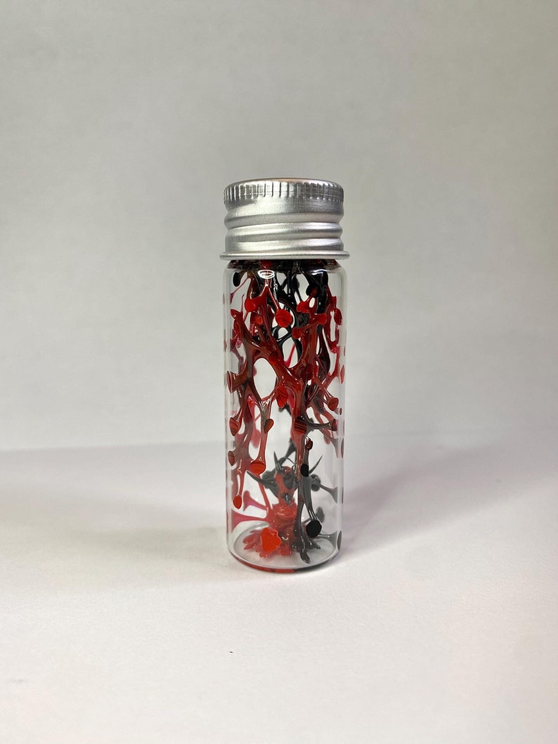 K&ouml;nnte beinhalten: Eine kleine Glasflasche mit silbernem Deckel, gef&uuml;llt mit roten und schwarzen Farbstichen.