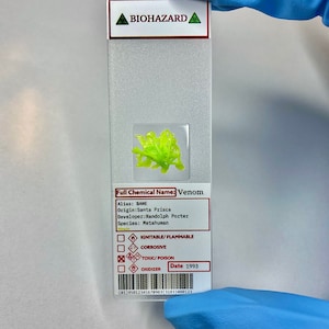 May include: A close-up of a microscope slide labeled "Venom" with a green substance inside. The slide also includes information about the substance, including its alias, origin, developer, species, and hazard warnings. The date "1993" is printed on the slide.