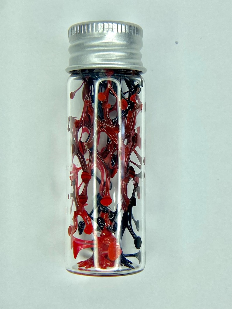 K&ouml;nnte beinhalten: Eine kleine Glasflasche mit silbernem Deckel, gef&uuml;llt mit roten und schwarzen Plastikteilen.