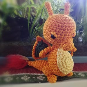 May include: Orange and green crocheted stuffed toy dragon with a red tail. The dragon has a yellow belly and black eyes.