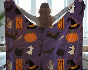 May include: A purple Halloween-themed blanket featuring a repeating pattern of pumpkins, witch hats, bats, ghosts, and the number 31. The blanket is held up, showcasing the design.