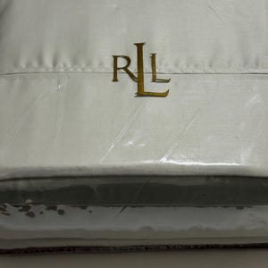 May include: A rectangular, light gray fabric item encased in clear plastic packaging. The center of the item features the gold-colored monogram letters RL in a serif font. The fabric appears smooth and folded, with a horizontal seam visible.