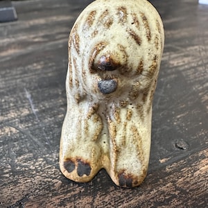 May include: A ceramic figurine of a dog, standing on all fours. The dog is white with brown markings and has a black nose and tongue. The figurine is glazed and has a rustic look.