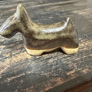 May include: A ceramic figurine of a dog in a brown and green glaze. The dog is standing on a wooden surface.