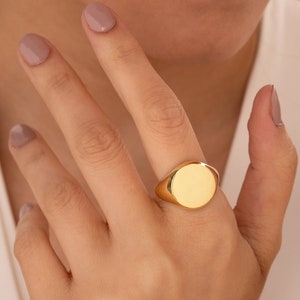 May include: A gold signet ring with a smooth, oval-shaped top. The ring is worn on the index finger of a hand.