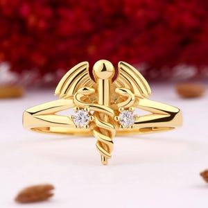 May include: A gold ring with a medical caduceus symbol and two small clear stones.
