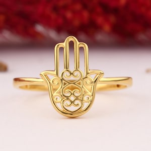 May include: A gold-colored ring featuring a Hamsa hand design. The ring's band is thin and simple, while the Hamsa hand has intricate, openwork detailing. The background is blurred, with hints of red and white.