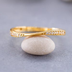 Op de afbeelding: Een goudkleurige ring met een zachte, golvende curve en een reeks kleine, heldere, fonkelende steentjes die langs de bovenkant van de band zijn ingelegd. Het metaal heeft een gepolijste afwerking die het licht over de structuur weerkaatst.