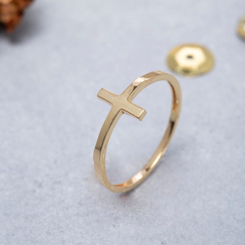 May include: A gold-toned ring featuring a simple cross design. The cross is positioned on the band of the ring, creating a minimalist and elegant look.