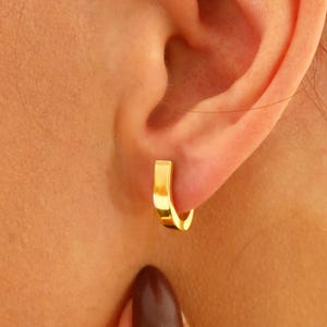 Könnte beinhalten: Nahaufnahme eines goldfarbenen quadratischen Creolen-Ohrrings. Der Ohrring wird am Ohr getragen. Der Ohrring hat eine glatte, reflektierende Oberfläche. Der Ohrring ist ein schlichtes, modernes Design.