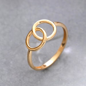 Op de afbeelding: Een gepolijste gouden ring met twee in elkaar grijpende cirkelvormige elementen in het midden. De band is glad en dun en sluit aan op de twee verbonden ringen die horizontaal aan de bovenkant liggen. Het metaal heeft een heldere afwerking.