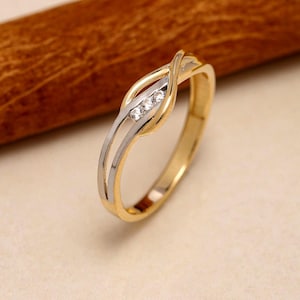 Könnte beinhalten: Ein zweifarbiger Ring mit einer polierten Gelbgoldschiene, die sich in ein überkreuztes Design teilt. Der Mittelteil ist mit einer silberfarbenen Einlage akzentuiert, die drei kleine, klare, rund geschliffene Steine in einer Reihe hält.