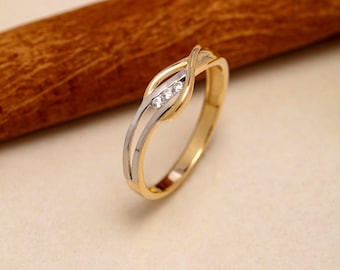 May include: A two-tone ring featuring a polished yellow gold band that splits into a crossover design. The center section is accented with a silver-toned inlay holding three small, clear, round-cut stones set in a vertical row along the band.