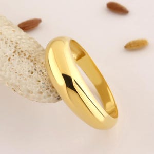 May include: A polished gold band ring with a smooth, rounded surface and a high-shine finish. The ring features a simple, solid construction with a uniform width throughout the entire circular band, reflecting light across its curved, metallic exterior.