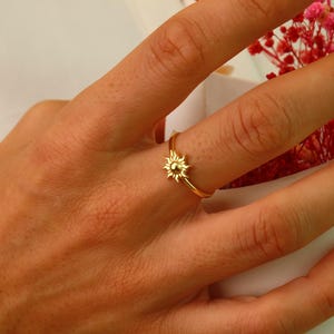 May include: A gold ring with a small sun design. The ring is on a person's finger.