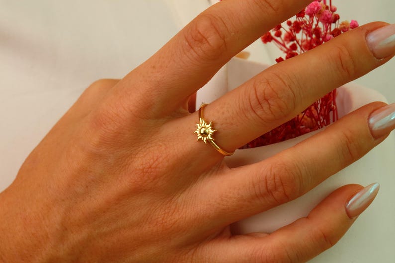 May include: A gold ring with a small sun design. The ring is on a person's finger.