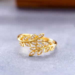 Könnte beinhalten: Ein goldfarbener Ring mit einem offenen Design, das zwei sich überlappende Blattzweige darstellt. Jedes Blatt ist mit kleinen, klaren, funkelnden Steinen besetzt, die das Licht einfangen und ein detailliertes botanisches Aussehen erzeugen.