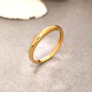 May include: A polished gold ring with a smooth, rounded band and a continuous, seamless design. The metal surface reflects light, highlighting the curved profile and the uniform, circular shape of this simple, solid gold jewellery piece.